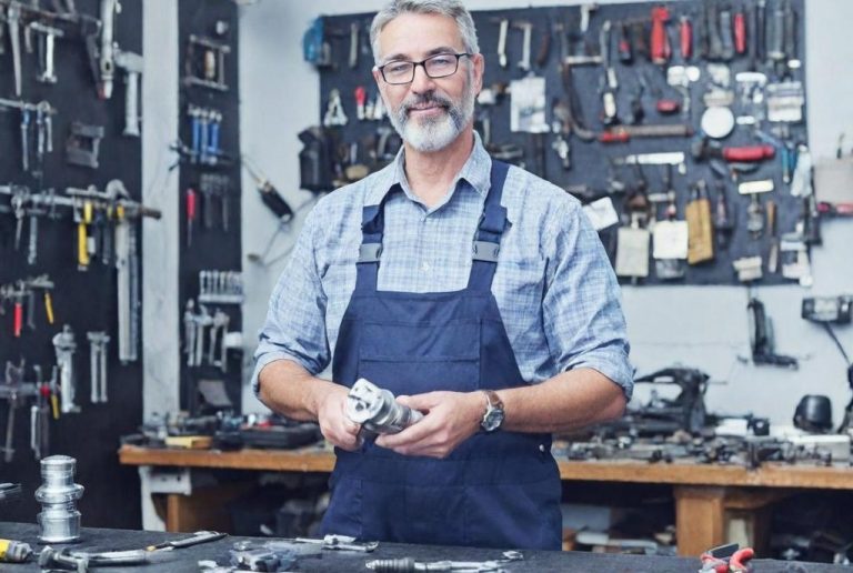 Mann in Latzhose steht in Werkstatt, hält Metallteile, Werkzeug an Wand.