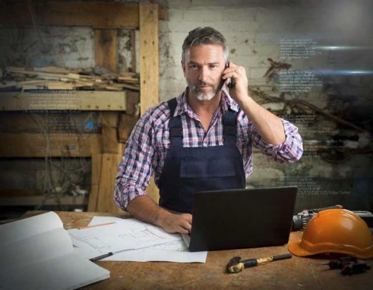 Ein Handwerker am Schreibtisch mit Laptop, Telefon und Bauplänen, in einer Werkstatt.
